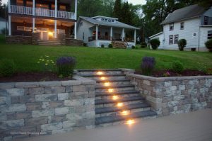 Stone steps with built-in lights lead up through a retaining wall to a grassy yard, with two porch-front houses in the background and flower beds on either side of the steps.