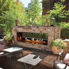An outdoor patio features a stone fireplace with visible flames, surrounded by potted plants and patio furniture. Sunlight filters through nearby trees, casting shadows on the table and ground.