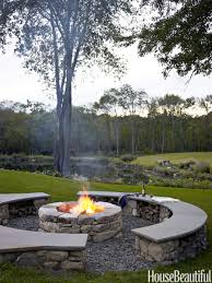 A circular stone fire pit with a burning fire is surrounded by curved stone benches on a grassy lawn, with trees and a scenic landscape in the background.