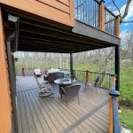 A view of a two-level outdoor deck with wood flooring and black railings; the lower deck has outdoor seating, a table, and overlooks a wooded area with leafless trees.