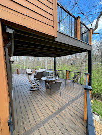 A view of a two-level outdoor deck with wood flooring and black railings; the lower deck has outdoor seating, a table, and overlooks a wooded area with leafless trees.
