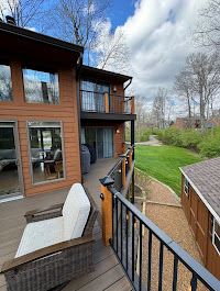 A view from a balcony with a wicker chair, overlooking a green yard, a garden shed, and a pathway. The brown house features large windows and a glass sliding door under a partly cloudy sky.