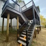 A two-level wooden deck attached to a house, with stairs featuring built-in lights, black railings, and an upper and lower covered seating area above a dirt yard.