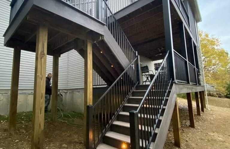 A two-level wooden deck attached to a house, with stairs featuring built-in lights, black railings, and an upper and lower covered seating area above a dirt yard.