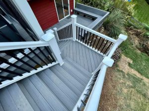 Wide-angle view of gray composite deck stairs with white posts and black balusters, attached to a red house, leading down toward a landscaped yard with grass, plants, and a built-in gray bench.