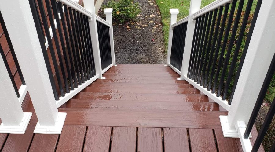 Wet wooden stairs with white and black railings lead from a deck down to a green lawn, bordered by mulch and small plants, next to a brick wall.
