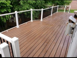 A freshly cleaned, brown wooden deck with white and black railing panels, surrounded by green trees and a brick building in the background.