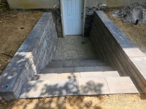 A stairway made of concrete blocks leads down between two retaining walls to a white door at the base of a building. The surrounding ground is bare soil.