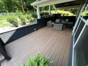 A modern outdoor patio features brown composite decking, black stone half-walls, and a sitting area with wicker chairs and a table, surrounded by greenery and partially covered by a roof.