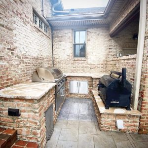 Outdoor brick kitchen with built-in grill, stainless steel cabinets, and a black smoker oven on stone countertops, located in a corner with brick walls and gray tiled flooring.