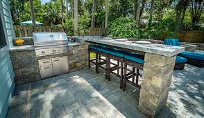 Outdoor patio with a built-in stainless steel grill, stone countertops, and a bar area with high stools. There is a seating area with cushions and trees in the background, creating a cozy outdoor kitchen space.