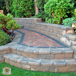 Curved stone steps lead up to a walkway made of rectangular pavers, bordered by low stone retaining walls, with green plants and bushes on either side in a landscaped garden.
