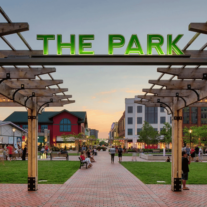 A large sign reading “THE PARK” in green letters marks the entrance to a lively outdoor plaza with people sitting on benches, walking, and buildings lining the area at sunset.