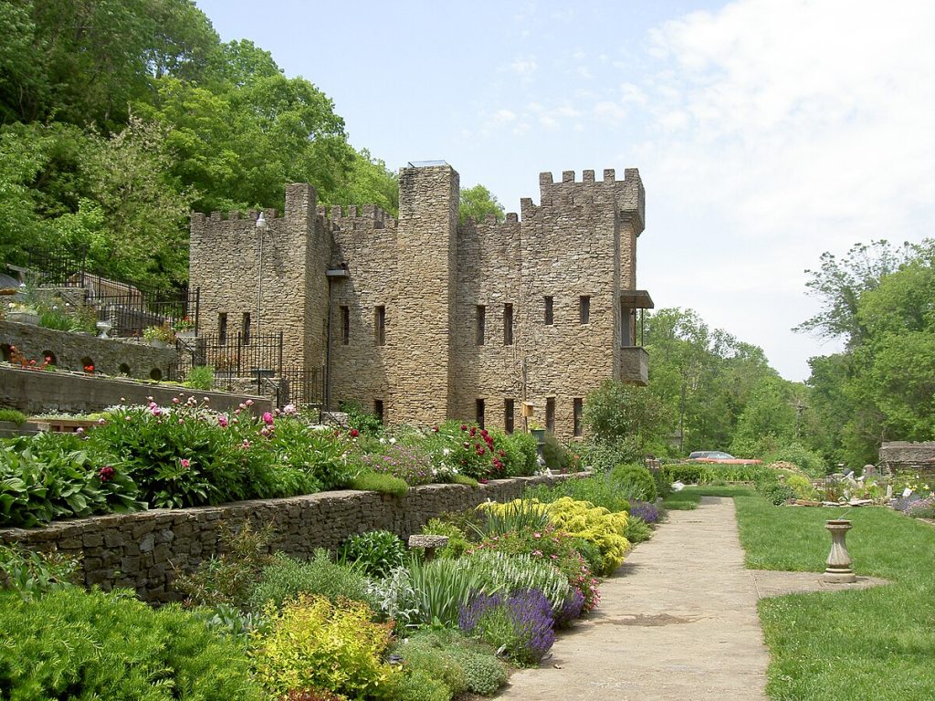 A stone castle with crenellated towers stands beside lush, terraced gardens filled with colorful flowers and greenery under a partly cloudy sky. A stone path runs alongside the garden toward the castle.