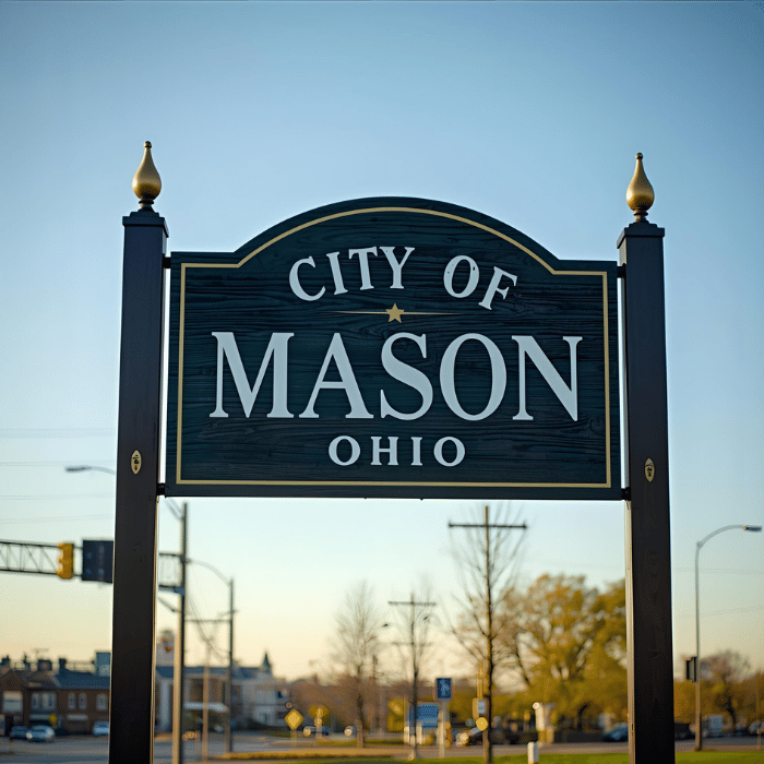 A sign reading City of Mason Ohio stands in the foreground, with a blurred view of a street, buildings, and leafless trees in the background under a clear sky.