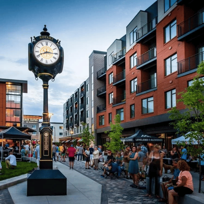 A lively street scene with people walking and gathering near a black vintage-style clock. Modern brick and glass apartment buildings line the street, and tents are set up for an outdoor event. The sky is partly cloudy. loveland ohio