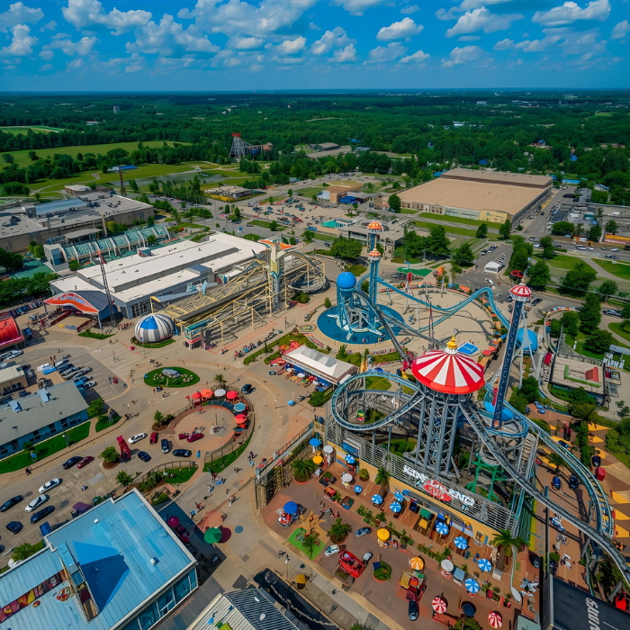 Aerial view of an amusement park with colorful rides, roller coasters, food stands, and outdoor seating, surrounded by green trees, roads, and neighboring buildings under a bright blue sky with scattered clouds. kings island graphic mason ohio