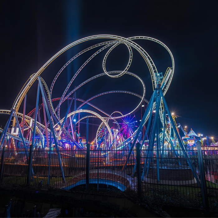 A brightly lit roller coaster with looping tracks glows against the night sky at an amusement park, colorful lights illuminating the ride and surrounding attractions beyond a dark metal fence. liberty township ohio