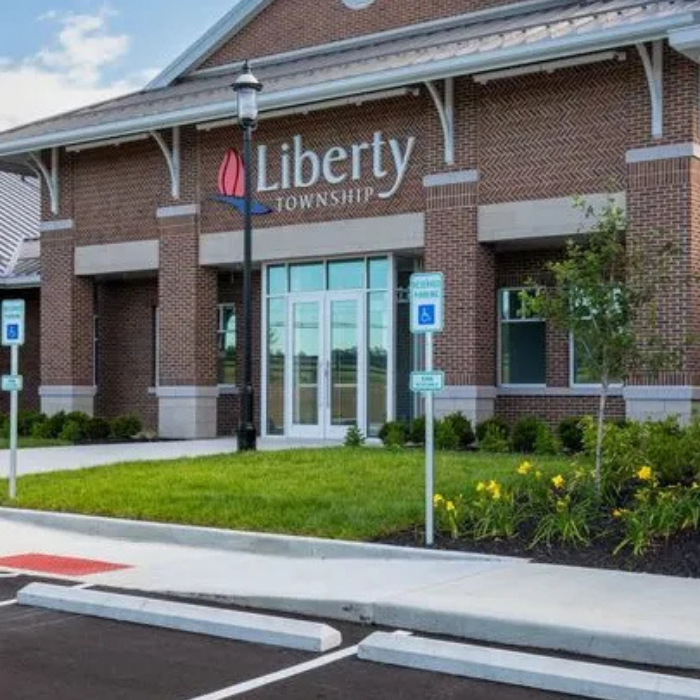 A brick building with large windows and a sign reading Liberty Township above the entrance. There are handicap parking spaces, signs, a lamp post, and landscaped greenery in front.