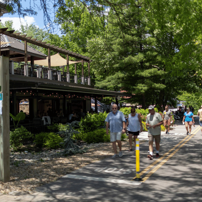 People walk and jog on a sunny, tree-lined paved trail beside a café with outdoor seating. The area is lush with greenery and has a relaxed, summery atmosphere.