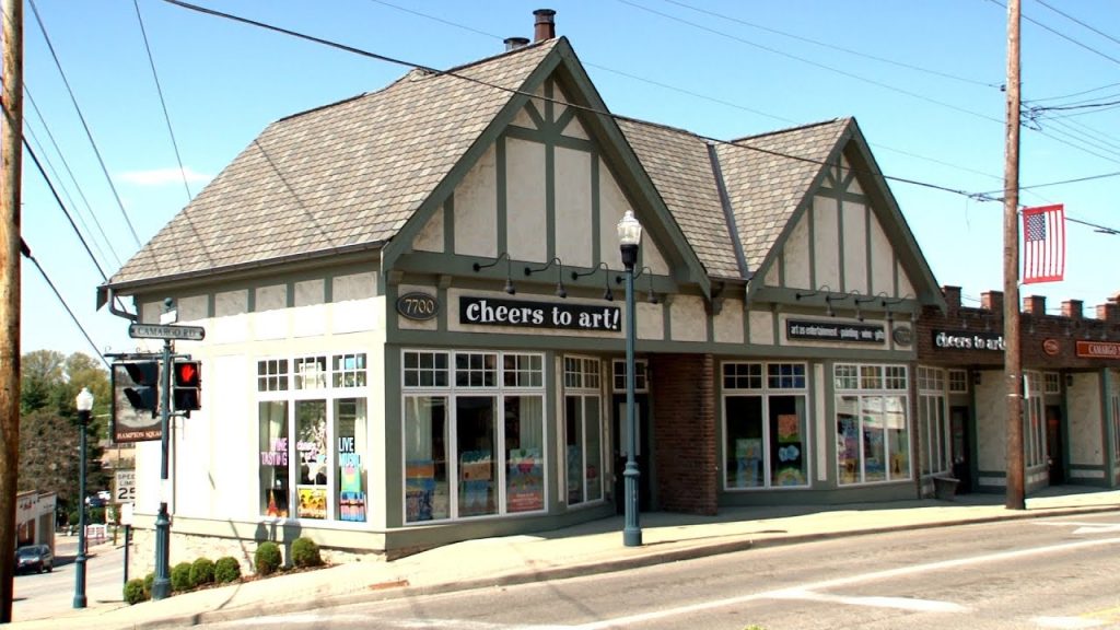 A corner building with Tudor-style architecture houses Cheers to Art! Large windows display colorful art supplies and posters. A streetlamp and traffic light stand outside, and an American flag is visible in the background.