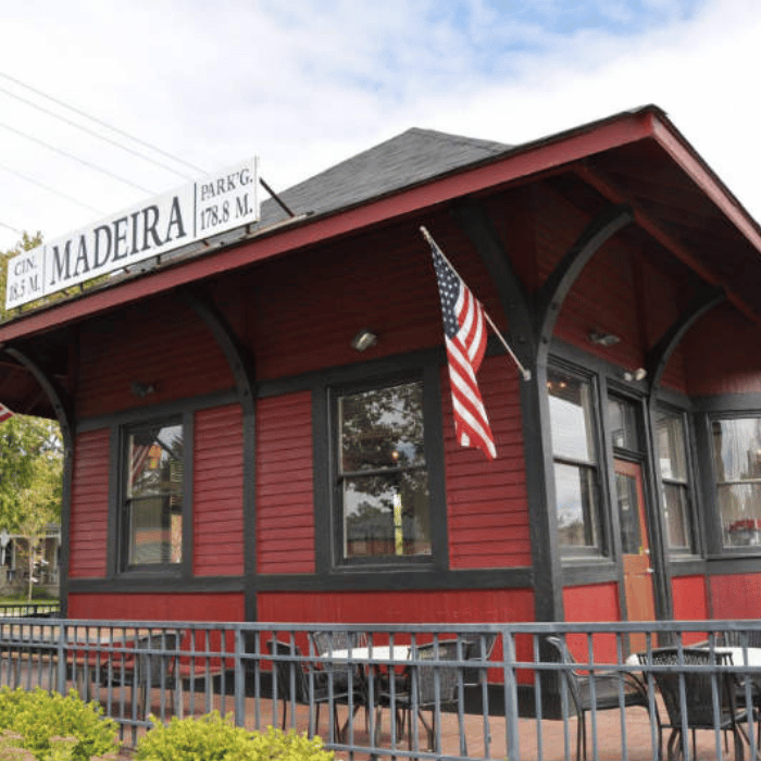 A historic red and black building with a sign reading MADEIRA, flying a U.S. flag, and surrounded by a black metal railing under a partly cloudy sky.