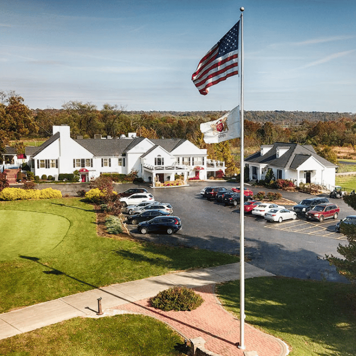 A large American flag waves in front of a white clubhouse with a parking lot full of cars, surrounded by green lawns and autumn trees under a clear sky. terrace park country club