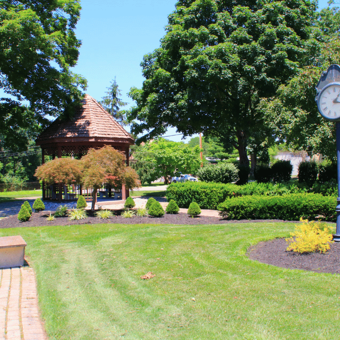 A landscaped park with a wooden gazebo, manicured shrubs, and a black street clock on a sunny day. The area is surrounded by lush green trees and well-kept grass. terrace park ohio