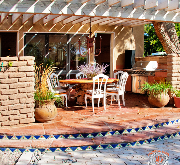 A covered outdoor patio with a wooden pergola, dining table, white chairs, potted plants, and a barbecue grill. Decorative tile borders the brick flooring, and sunlight creates shadows on the seating area.