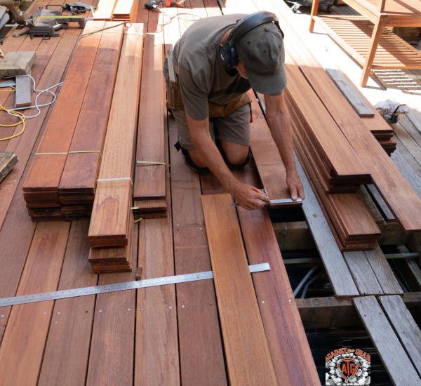 A man wearing headphones and work clothes measures wooden planks while kneeling on a partially constructed wooden deck, surrounded by stacked wood and tools.