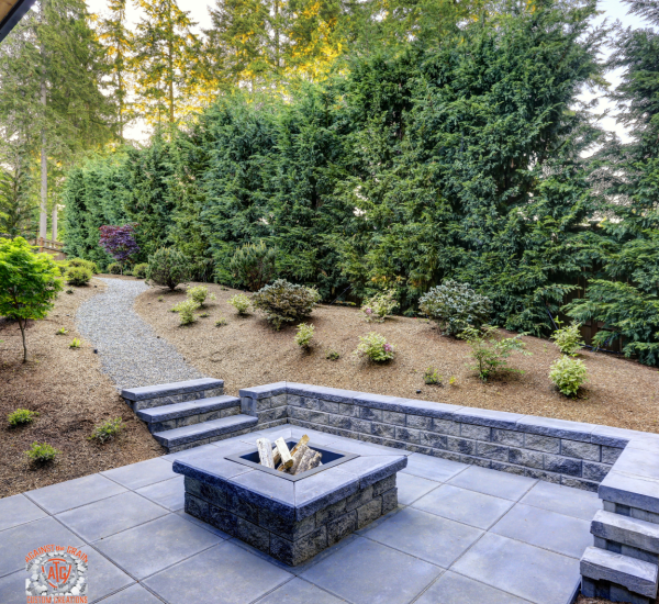 A modern stone patio with a square fire pit and surrounding pavers overlooks landscaped beds, shrubs, and a gravel path leading up stone steps, bordered by tall evergreen trees.