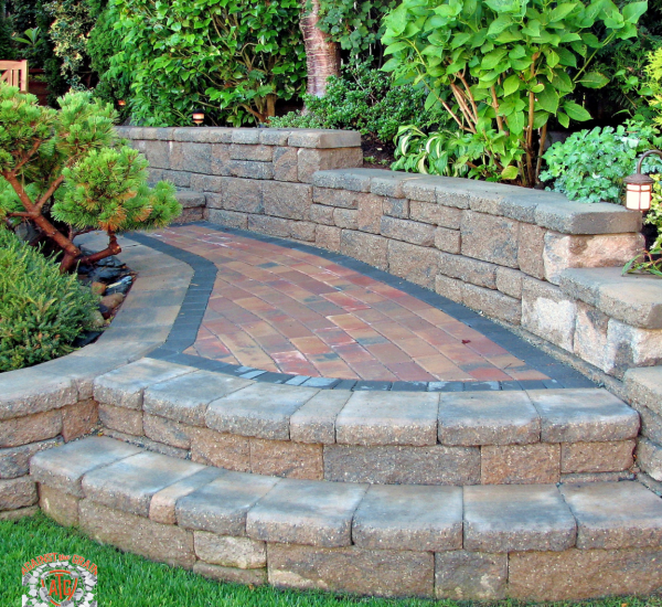 A curved brick pathway with two wide steps and raised stone walls, surrounded by lush green plants and shrubs in a well-maintained garden.