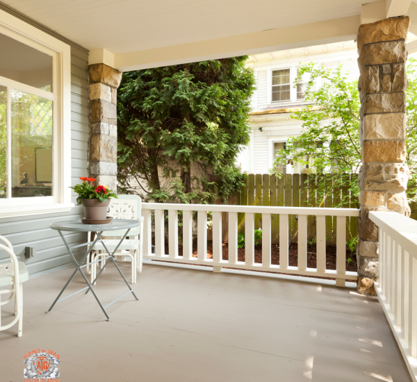 A cozy front porch with white railings, stone pillars, and a small round table with two chairs. A potted plant sits on the table. Trees and a wooden fence are visible in the background.